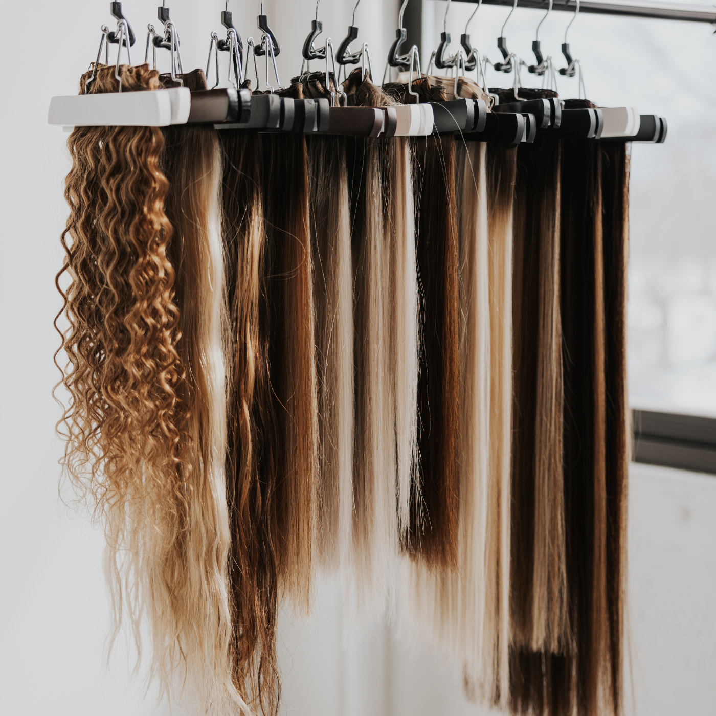 Various shades and textures of hair extensions hanging neatly on white hangers, representing exclusive pro stylist access.