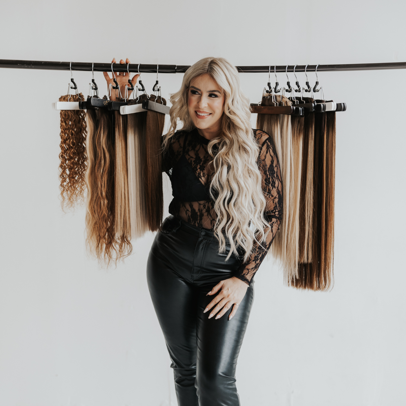 Blonde woman standing in front of hanging hair extension wefts, smiling and wearing black lace top and leather pants